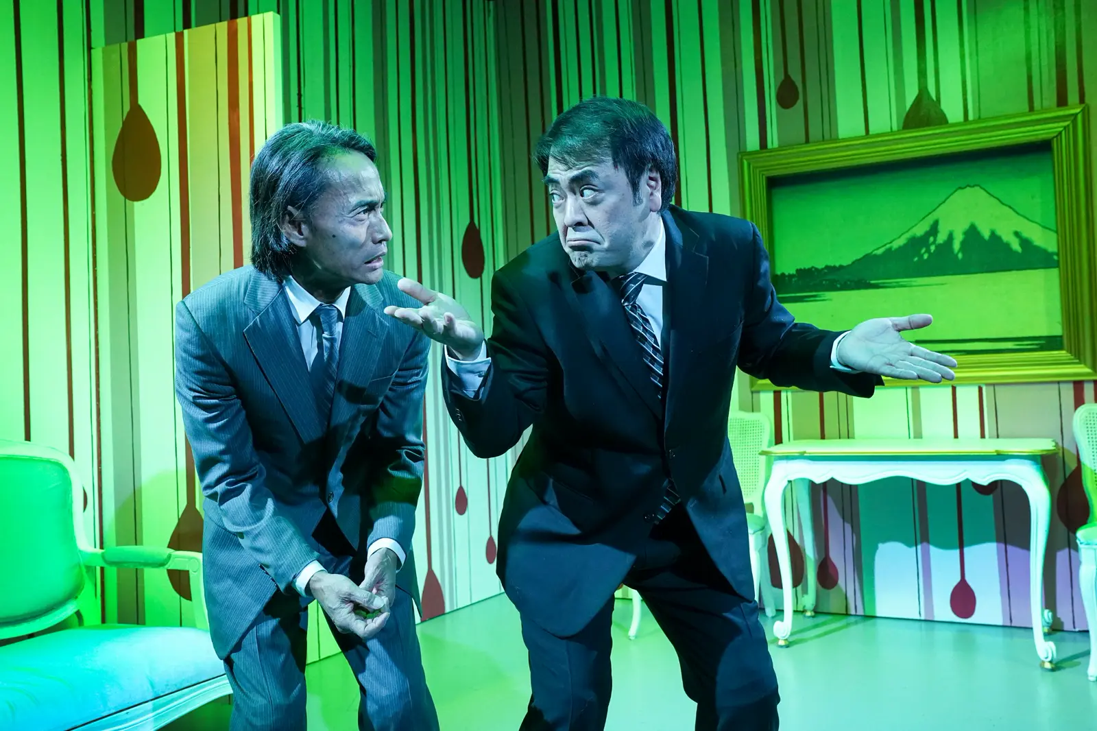 Two actors in suits on a green-lit theater set